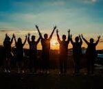 A diverse group of friends raises their arms in celebration against a vibrant sunset backdrop.